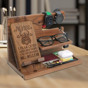May include: A wooden phone stand with a personalized message that reads "I'm a man of the Norse. I fear Odin and my wife. You are neither Mr. William". The stand has slots for a phone, watch, glasses, and other accessories.