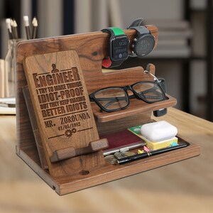 May include: A wooden desk organizer with multiple compartments. It features a phone holder, watch holders, a glasses tray, and a small tray for accessories. The organizer has the text "ENGINEER" and a quote engraved on the front.