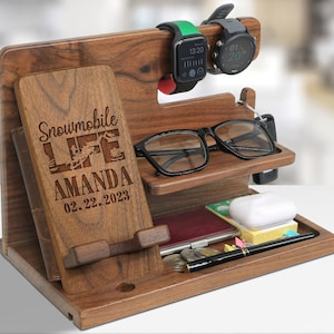 May include: A wooden desk organizer with multiple compartments. It features a phone holder with the text "Snowmobile LIFE AMANDA 02.22.2023", a shelf for glasses, and a tray for small items. Two smartwatches are displayed on top.