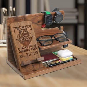 May include: A wooden desk organizer with multiple compartments and slots. It features a slot for a phone or tablet with the text "THAT WHICH DOES NOT KILL ME SHOULD RUN MR. WILLIAM". It holds glasses, watches, keys, and other small items.