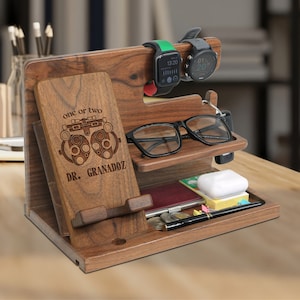 May include: A wooden organizer with a phone stand, watch holder, glasses holder, and a small compartment for keys or other items. The organizer is engraved with the text "one or two" and "DR. GRANADOZ" and features a stylized eye chart.