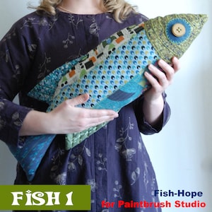 Könnte beinhalten: Ein handgefertigtes Patchwork-Fischkissen in Grün, Blau und Gelb. Das Kissen hat ein Knopfauge und wird von einer Person in einem lila Shirt gehalten. Der Text "FISH 1" ist in Grün auf grünem Hintergrund. Der Text "Fish-Hope for Paintbrush Studio" ist in Rot und Weiß.