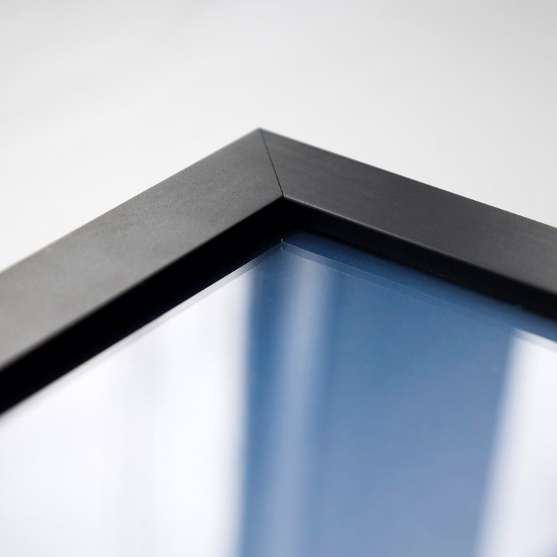 May include: Close-up of the corner of a black picture frame with a clear glass insert. The frame is made of wood and has a smooth finish.