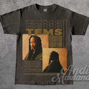 May include: Dark gray t-shirt featuring the word "TEMS" in gold lettering. The shirt has two images of a person with long braids. The text "FOR BROKEN EARS" and a barcode are also visible. The shirt is part of the "MERCH" collection.