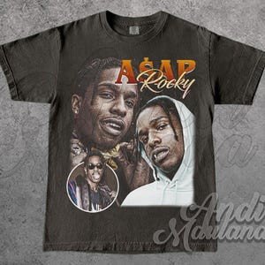 Puede incluir: Camiseta gris oscuro con múltiples imágenes de A$AP Rocky. La camiseta tiene un diseño vintage con el nombre "A$AP Rocky" en letras naranjas y doradas. También se incluye una imagen circular de A$AP Rocky con gafas de sol.