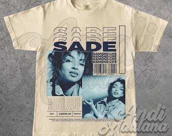May include: Cream-colored t-shirt featuring a blue-toned image of a woman and the name "SADE" in a layered design. The shirt includes the text "DIAMOND LIFE" and a barcode, with additional text at the bottom.