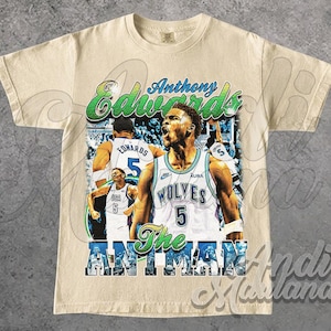 Puede incluir: Camiseta color crema con un gráfico del jugador de baloncesto Anthony Edwards. El diseño incluye el texto "Anthony Edwards" en verde y azul, junto con "Wolves" y "The Ant Man".