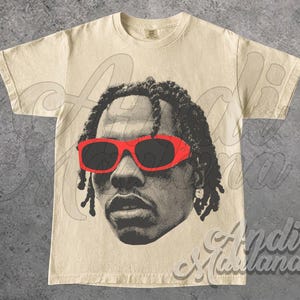 May include: Cream-colored t-shirt featuring a halftone portrait of a person wearing red sunglasses. The graphic is in black and gray tones. The shirt has a crew neck and short sleeves.