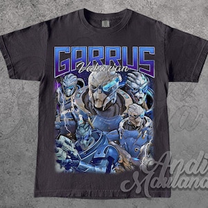 Puede incluir: Camiseta gris oscuro con un diseño gráfico del personaje Garrus Vakarian de la serie Mass Effect. El diseño incluye el nombre "GARRUS" en texto azul y morado, junto con múltiples imágenes del personaje con armadura azul y gris.