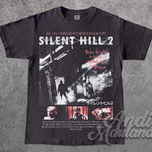 May include: Dark gray t-shirt featuring a graphic from Silent Hill 2. The design includes Japanese text, the game's title, and imagery of figures in a dark, urban setting. The shirt also has text that reads "The door that wakes in darkness, opening nightmare."