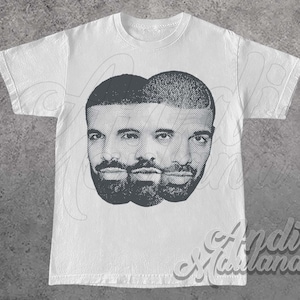 Może przedstawiać: Biała koszulka z szarą grafiką twarzy powtórzoną trzykrotnie. Projekt ma efekt półtonów. Koszulka ma okrągły dekolt i krótkie rękawy. Podpis artysty, "Andi Marlana", jest widoczny w prawym dolnym rogu.