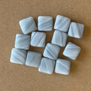 May include: A collection of twelve square, light blue glass tiles with a swirled, marbled pattern. The tiles are arranged on a brown surface. Each tile has rounded corners and a glossy finish, suitable for mosaic projects or decorative use.