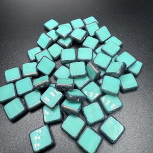 May include: A collection of turquoise square beads with a metallic dark blue border. The beads are scattered, showcasing their glossy surface and diamond shape. Ideal for jewelry making or crafting projects.