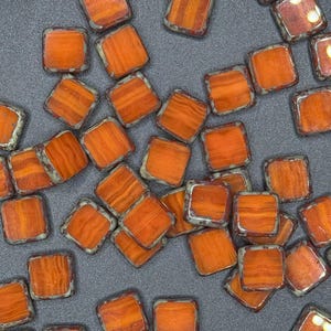 May include: A collection of square beads with an orange, wood-grain appearance. Each bead has a silver-colored border. The beads are scattered on a gray surface. These beads are suitable for jewelry making and crafting.