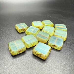 May include: A collection of square glass beads in shades of yellow, green, and blue. Each bead has a slightly irregular shape and a textured surface. The beads are scattered on a dark surface, creating a visually appealing arrangement.