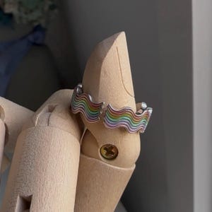 May include: A silver ring with a rainbow wave design. The ring is on a wooden ring display stand.