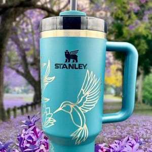 Peut inclure: Une tasse de voyage en acier inoxydable bleu turquoise avec une poign&eacute;e noire et un motif d'oiseau-mouche dor&eacute;. La tasse a un couvercle noir avec un trou pour paille et le logo Stanley est imprim&eacute; sur le devant.
