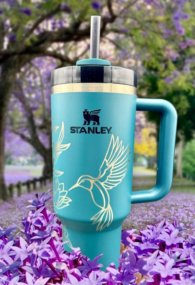 Peut inclure: Une tasse de voyage en acier inoxydable bleu turquoise avec une poign&eacute;e noire et un motif d'oiseau-mouche dor&eacute;. La tasse a un couvercle noir avec un trou pour paille et le logo Stanley est imprim&eacute; sur le devant.