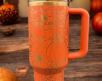 May include: An orange Stanley tumbler with a gold-toned floral and pumpkin line art pattern. The cup features a matching orange handle, a clear lid with a rotating closure, and a clear straw. The text Pumpkin Spice is printed at the bottom.