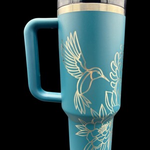 Peut inclure: Mug de voyage bleu sarcelle avec un couvercle en acier inoxydable et une poign&eacute;e. Le mug est orn&eacute; d'un motif grav&eacute; au laser d'un colibri et de fleurs.