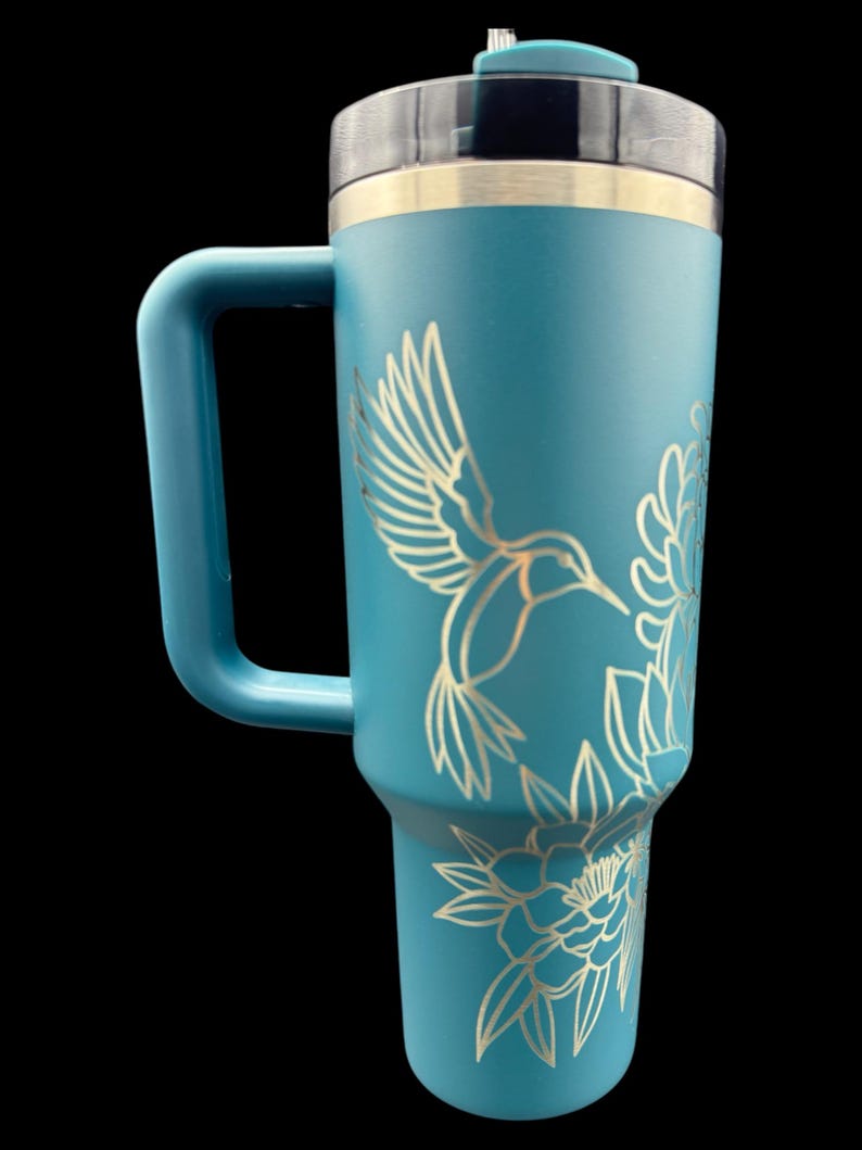 Peut inclure: Mug de voyage bleu sarcelle avec un couvercle en acier inoxydable et une poign&eacute;e. Le mug est orn&eacute; d'un motif grav&eacute; au laser d'un colibri et de fleurs.
