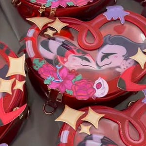 May include: Red heart-shaped purse with a clear front, featuring a cartoon couple and floral accents. The purse has a gold zipper, star-shaped embellishments, and a red handle. The design includes a red and black striped pattern.