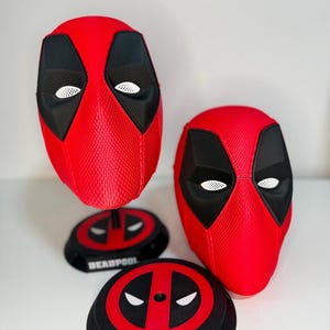 May include: Two red and black Deadpool character masks with textured surfaces and white mesh eye lenses. One mask is mounted on a black circular display stand featuring the Deadpool logo and the text DEADPOOL, while the other sits beside it.