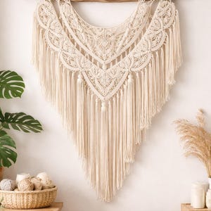 May include: Cream-colored macrame wall hanging with intricate geometric patterns and long fringe. The textile art piece is suspended from a natural wooden dowel, adding a touch of bohemian decor to any space. The wall hanging is a beautiful example of handcrafted textile art.