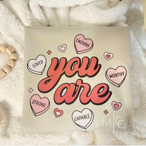 May include: Beige canvas pillow with the text "you are" in large red letters.  The pillow is decorated with pink and white heart-shaped candy with words like "loved", "strong", "capable", "worthy", and "enough".