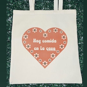 May include: A white canvas tote bag with a red heart design. The heart has white flowers around the edge and the text "Hay comida en la casa" in a retro font.
