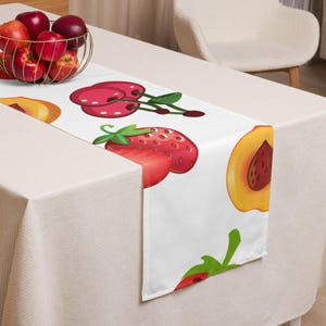 Puede incluir: Camino de mesa blanco con un patrón de frutas colorido. El diseño incluye ilustraciones de cerezas, fresas y melocotones. El camino de mesa está colocado sobre un mantel blanco, con una cesta de alambre con manzanas en el fondo.