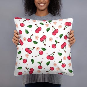 May include: White square pillow with a repeating pattern of red cherries, green leaves, and small pink flowers. The cherries have dark green stems and are scattered across the pillow's surface. The pillow is held by a person.