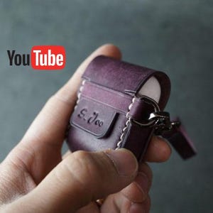 May include: A purple leather AirPod case with white stitching and a silver clasp. The case has a small flap with the embossed text "S. Joo". The YouTube logo is visible in the background.