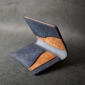 以下が含まれることがあります： 二つ折りデザインのオープンレザーウォレット。ウォレットは、タンとグレーのインテリアアクセントを備えた濃い青色の外観が特徴です。ウォレットは開いており、内部のカードスロットと対照的な色を示しています。