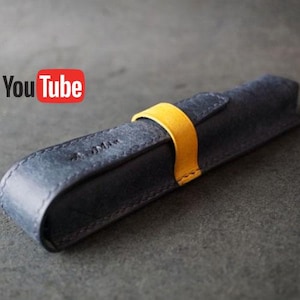 Op de afbeelding: Een blauwe leren etui met een gele bandsluiting. De etui heeft een gebogen ontwerp en een gestikte rand. Het YouTube-logo is zichtbaar in de linkerbovenhoek. De etui is waarschijnlijk ontworpen om één pen of potlood te bevatten.