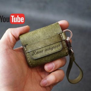 Op de afbeelding: Olijfgroene leren AirPods-hoes met een riem en sleutelhanger. De naam "Kwon sungoon" is op de voorkant in reliëf aangebracht. Het YouTube-logo staat op de achtergrond.
