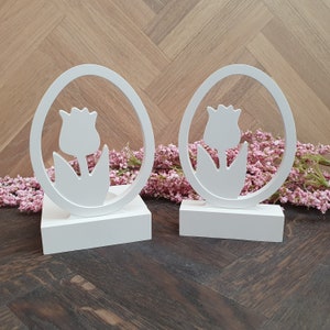 Könnte beinhalten: Zwei weiße, tulpenförmige Kerzenhalter aus Holz mit einem Ausschnittmuster. Die Halter stehen auf einer Holzoberfläche mit rosa Blumen.