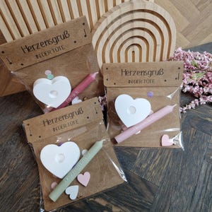 Könnte beinhalten: Mehrere Geschenksets in Klarsichttüten, jeweils mit einer weißen herzförmigen Dekoration, einer farbigen Kerze und Konfetti. Die Tüten haben einen braunen Papieraufsatz mit dem Text "Herzensgruß IN DER TUTE".