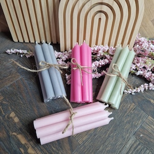 Könnte beinhalten: Bündel von Stabkerzen in verschiedenen Farben: Grau, Rosa und Grün. Jedes Bündel ist mit Bindfaden gebunden. Ein Holzbogen und rosa Blumen befinden sich im Hintergrund. Die Kerzen sind auf einer dunklen Holzoberfläche angeordnet.