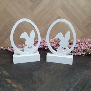 Könnte beinhalten: Zwei weiße, hölzerne Osterhasen-Eierhalter. Jeder Halter ist eiförmig und hat eine Hasensilhouette im Inneren. Der Hase hat ein kleines Herz auf der Brust. Die Halter stehen auf einer dunklen Holzoberfläche.