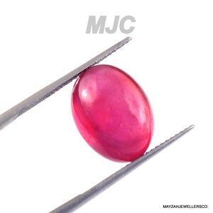 May include: A vivid, oval ruby gemstone held by tweezers. The gemstone is a deep, saturated red colour with a smooth, polished surface. The image includes the text "MJC" and "MAYZAHJEWELLERSCO."