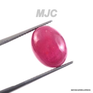 May include: A polished, oval-shaped ruby gemstone, held by tweezers against a white background. The ruby is a deep, vibrant red color, with a smooth, reflective surface. The text "MJC" is visible in the upper center.