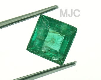 Puede incluir: Una esmeralda verde vibrante, de corte cuadrado, se sostiene con pinzas sobre un fondo blanco. La gema tiene características internas visibles y es de un color verde rico y saturado. El texto "MJC" y "MAYZAHJEWELLERSCO." son visibles.