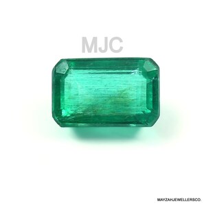 Può includere: Uno smeraldo rettangolare di colore verde vibrante. La gemma ha un taglio sfaccettato con caratteristiche interne visibili. Le lettere "MJC" sono sopra la gemma. Il testo "MAYZAHJEWELLERSCO." è sotto.