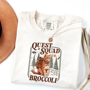 May include: Cream-colored t-shirt with a graphic featuring a cat with a backpack, the words "QUEST SQUAD" and "BROCCOLI". The design includes trees, birds, and the text "No One Wants It, But It's Good For You". A gold necklace and a brown hat are also visible.
