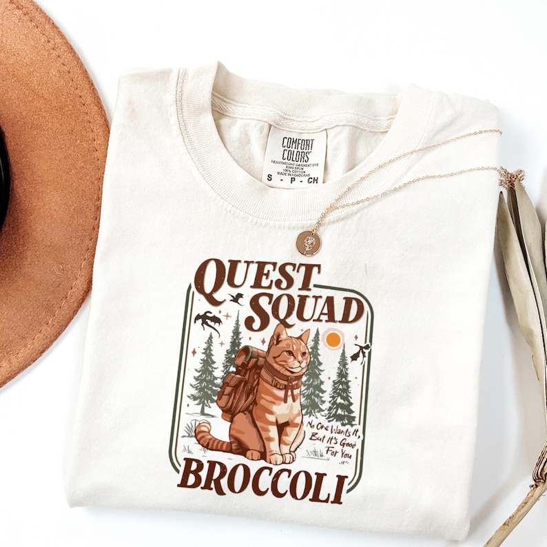 May include: Cream-colored t-shirt with a graphic featuring a cat with a backpack, the words "QUEST SQUAD" and "BROCCOLI". The design includes trees, birds, and the text "No One Wants It, But It's Good For You". A gold necklace and a brown hat are also visible.