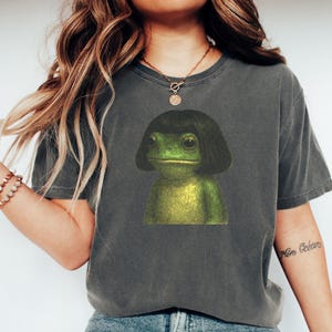 May include: Dark grey t-shirt featuring a whimsical illustration of a frog with a black bob haircut. The frog is green and yellow, with large eyes. The shirt is a casual style, suitable for everyday wear.