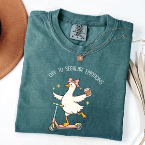 Op de afbeelding: Teal Comfort Colors T-shirt met een afbeelding van een eend op een scooter met een koffiekopje. De tekst "OFF TO REGULATE EMOTIONS" staat boven de eend gedrukt. Een gouden ketting ligt over het shirt.