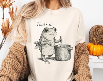 Op de afbeelding: Crèmekleurig T-shirt met een kikkerkok die in een pot roert, met een schort aan. De tekst "That's it." staat boven de kikker. Het shirt heeft een relaxte pasvorm.