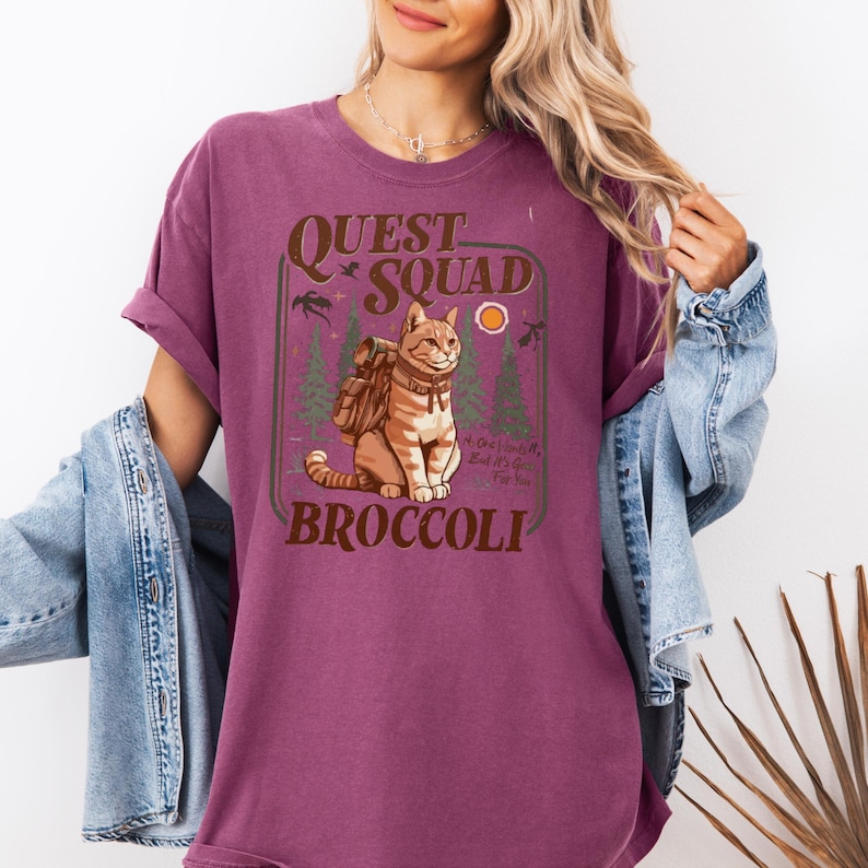 May include: A maroon t-shirt with a graphic featuring a cat wearing a backpack, the words "Quest Squad" and "Broccoli". The design includes trees, birds, and a sun. The shirt has rolled-up sleeves.